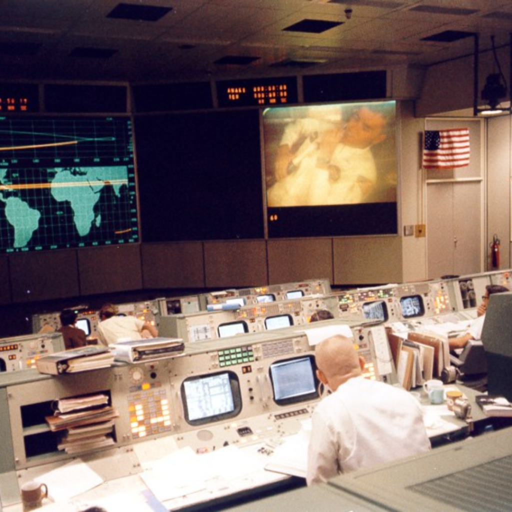 Gene Kranz, with his back to us, at right center watches a TV transmission from the Apollo 13 crew moments before the accident that crippled the mission. Fred Haise can be seen on the large screen at the upper right. 13 April 1970. Scan by Kipp Teague.